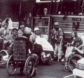 Archival photograph from Smithsonian exhibit on the history of the ADA. Veterans in wheelchairs gathered at a bus station. One has a sign on the back saying “I can’t even get to the back of the bus”